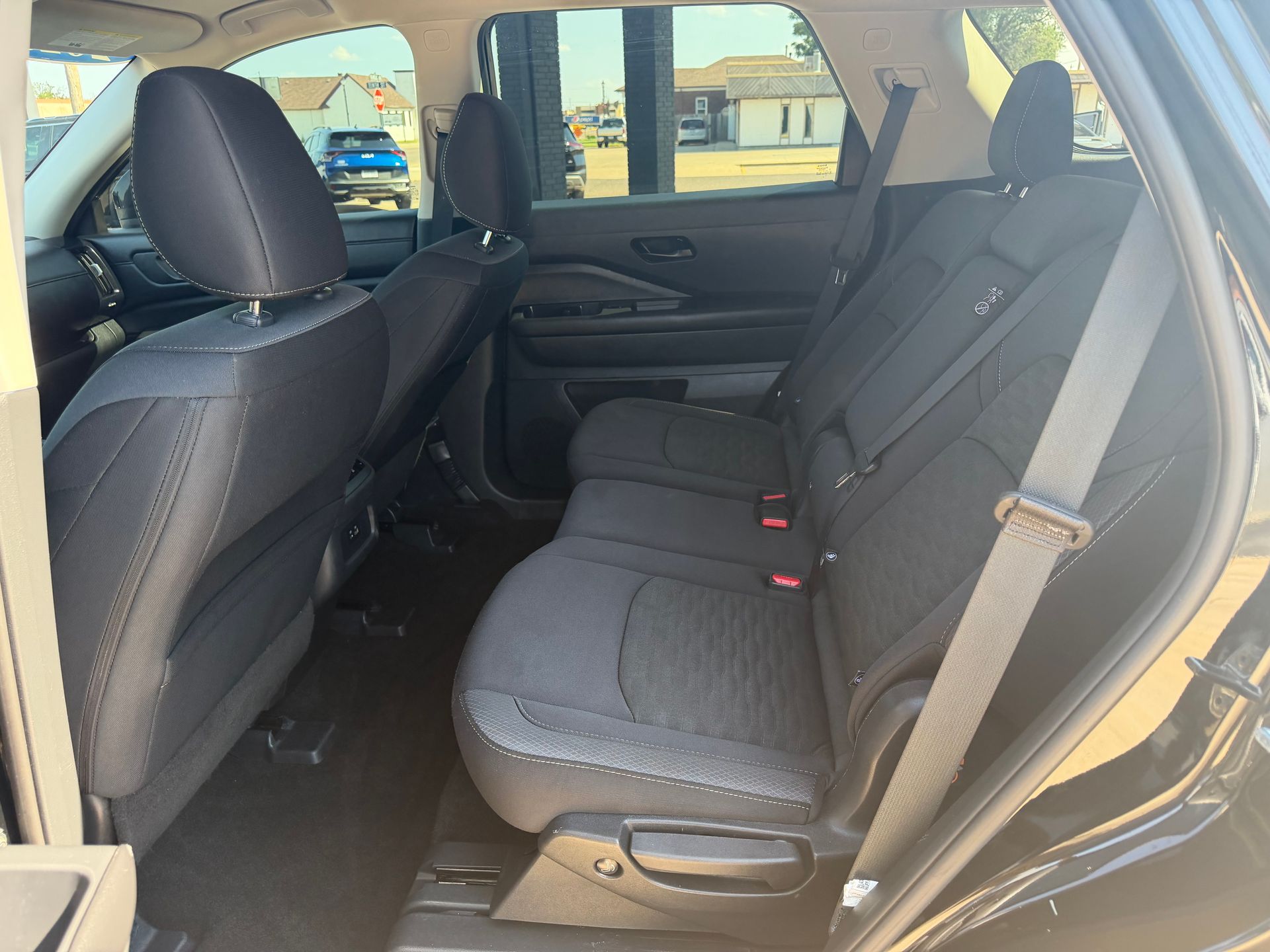 Empty rear seat of a car with black upholstery and beige seatbelts, viewed from the open door