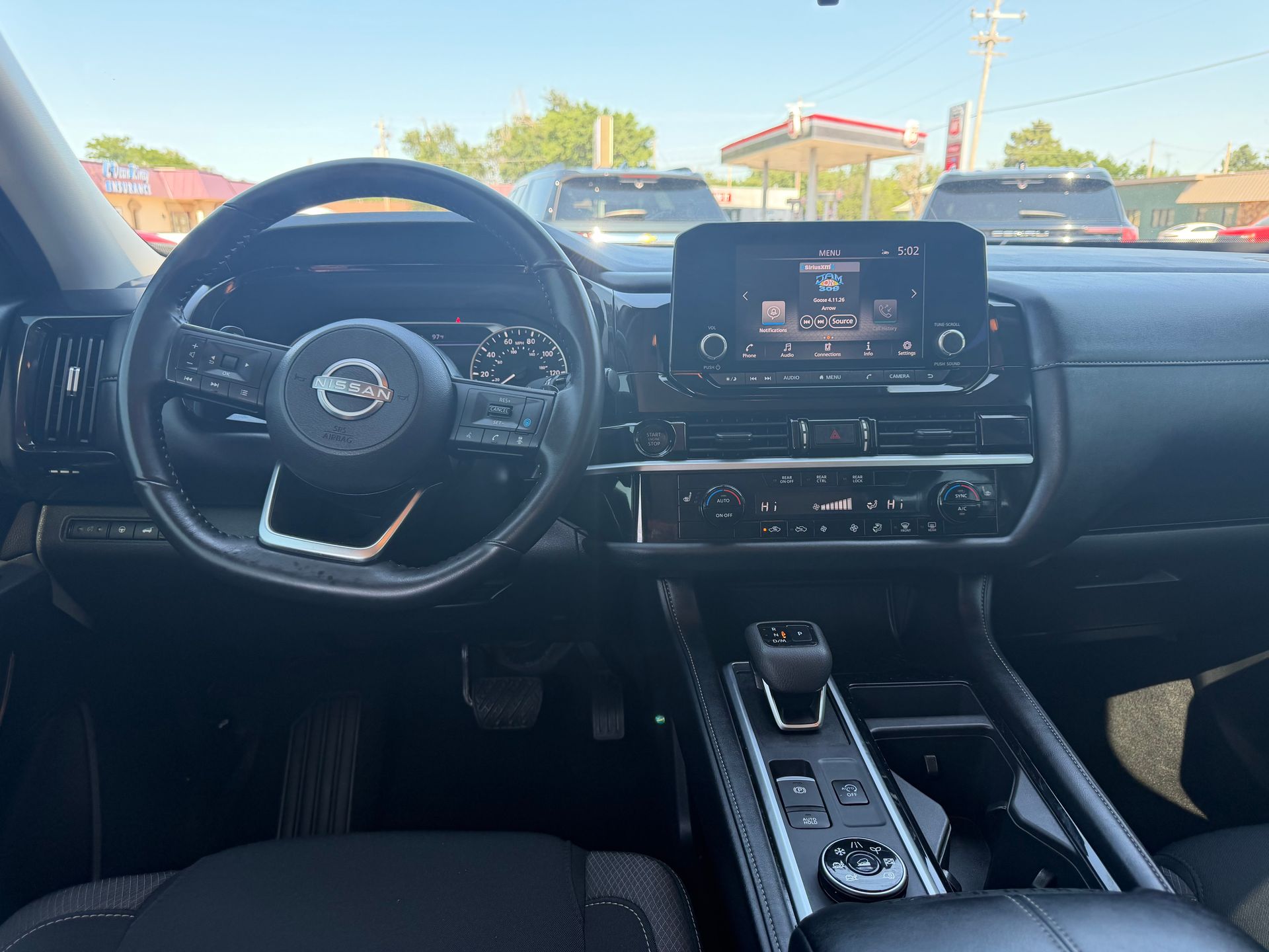 Car interior view of a Nissan dashboard, steering wheel, center touchscreen, and gear shifter in daylight