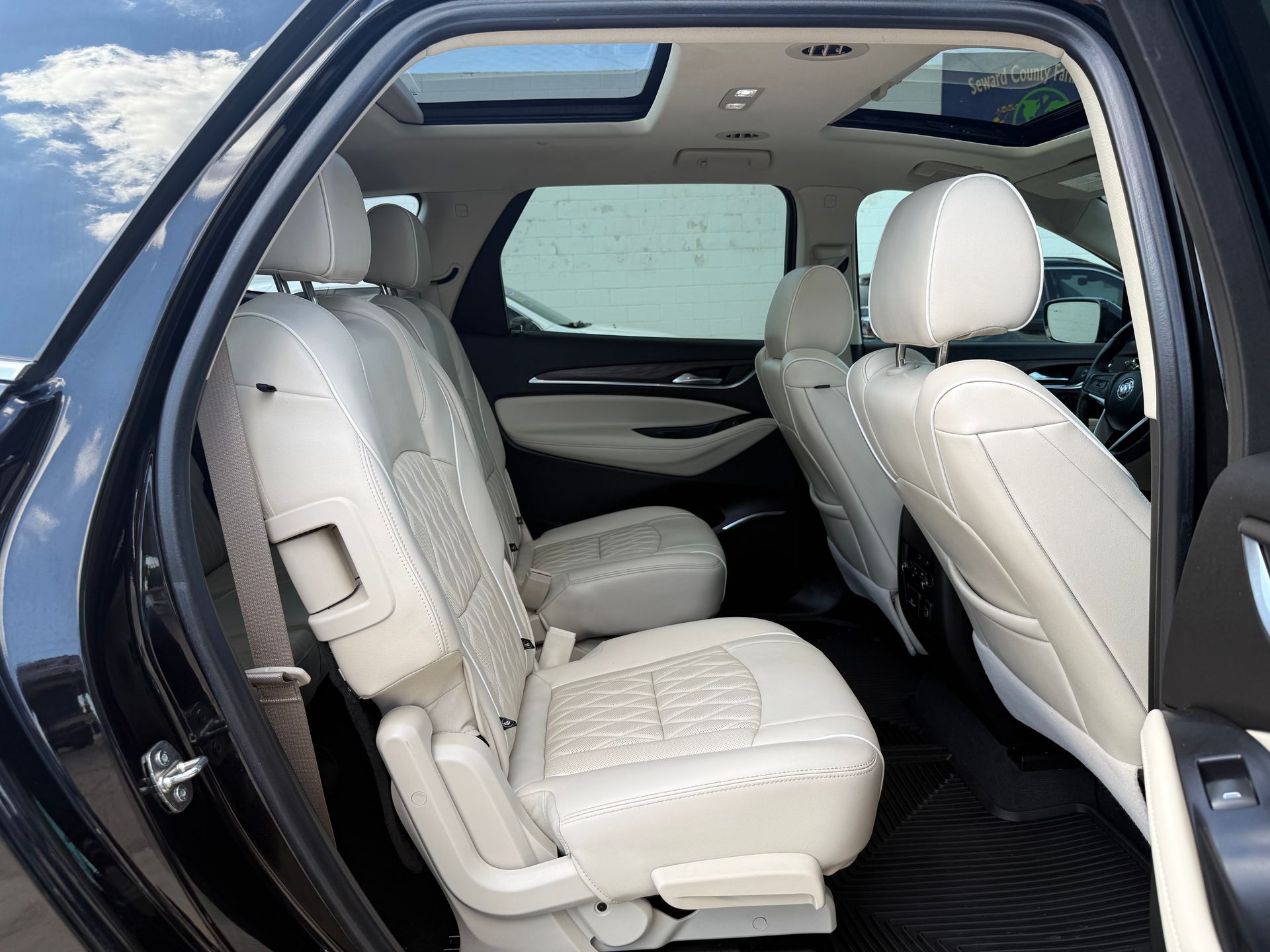 Interior view of a vehicle: cream leather seats, open door, sunroof, gray carpet, sunny outdoors.