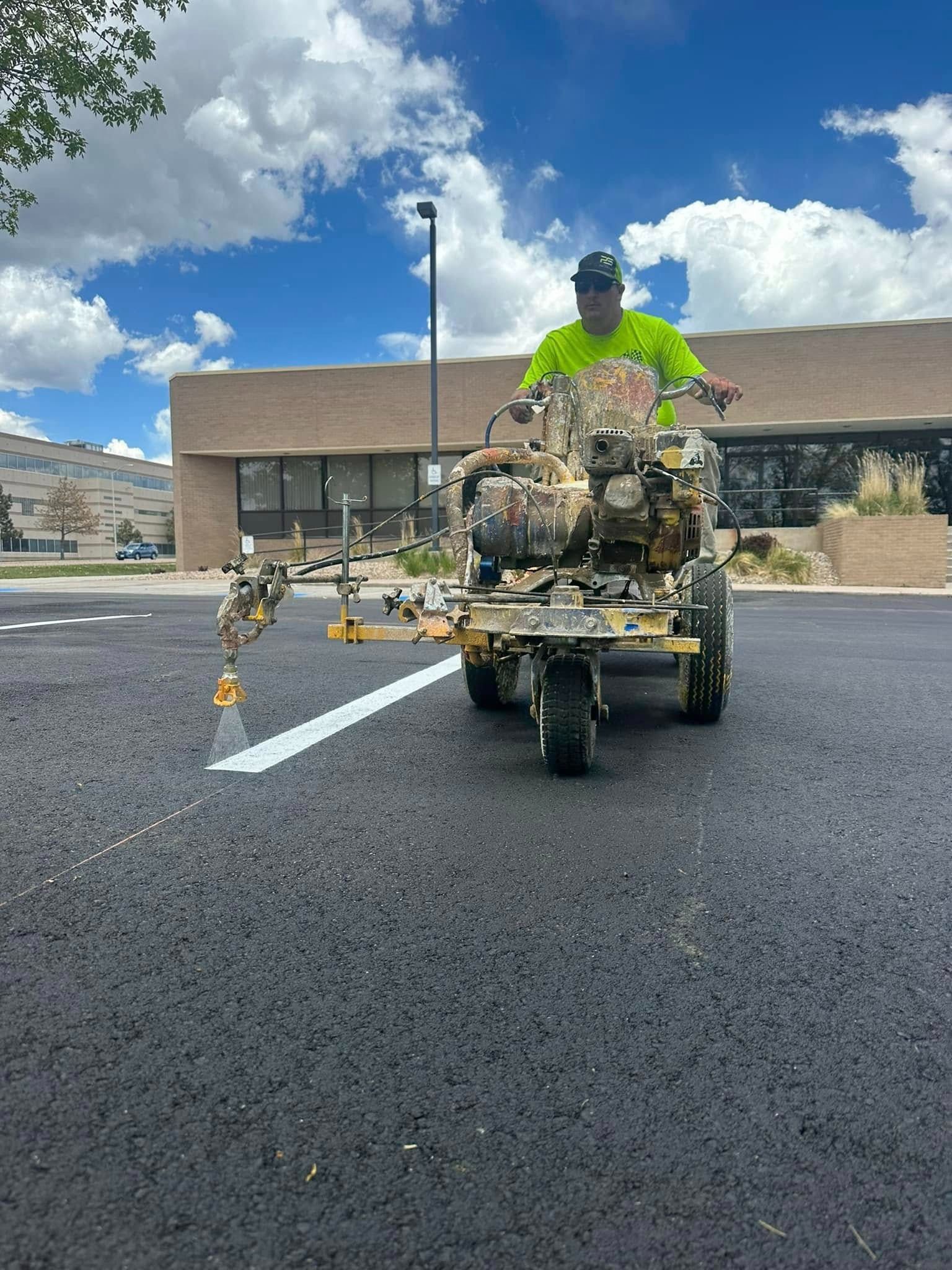 Person operating a road marking machine, painting a white line on asphalt. Bright sun, building in background.