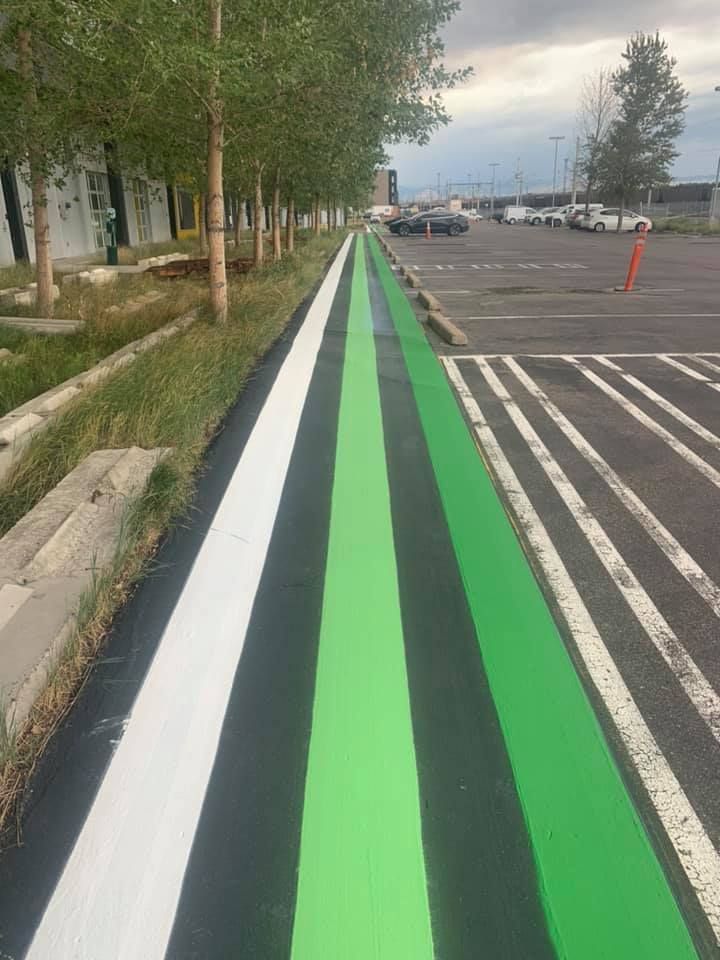 Striped path painted in white, black, and green, alongside a parking lot and trees.