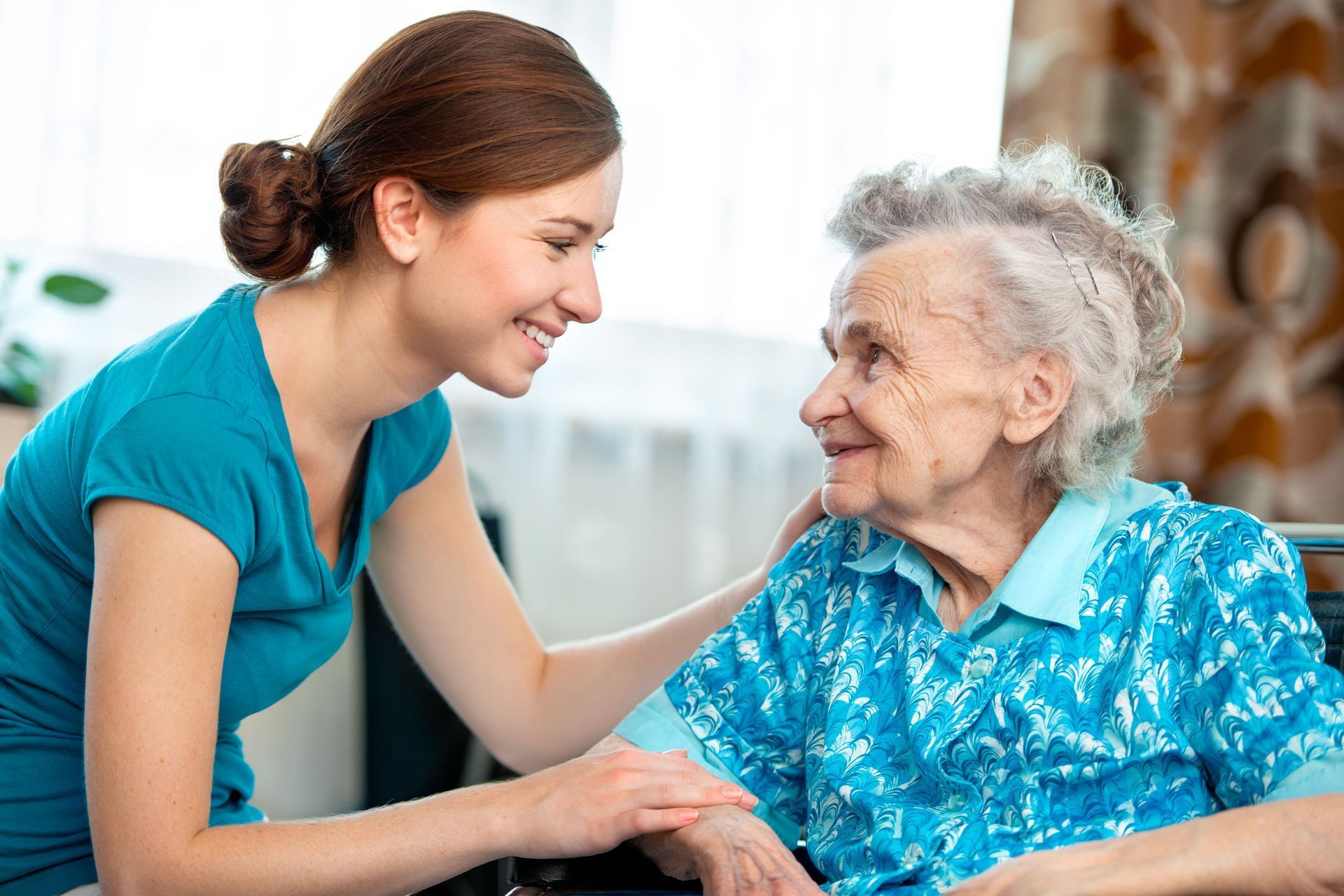 Caregiver in teal comforting an elderly woman in a blue blouse indoors, sharing a warm smile