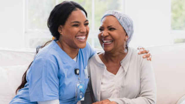 Two smiling women sitting on a couch, one in blue scrubs, sharing a happy moment