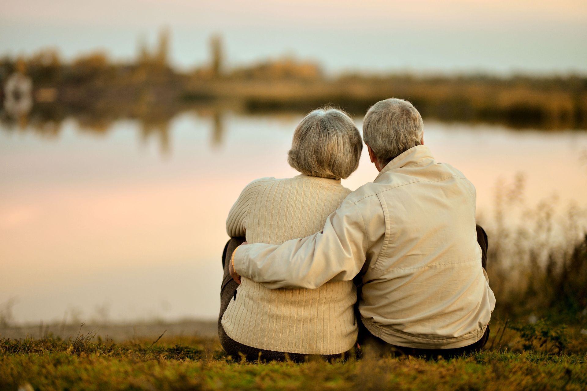 Two people sit on a grassy bank, embracing and looking over a calm lake at sunset.