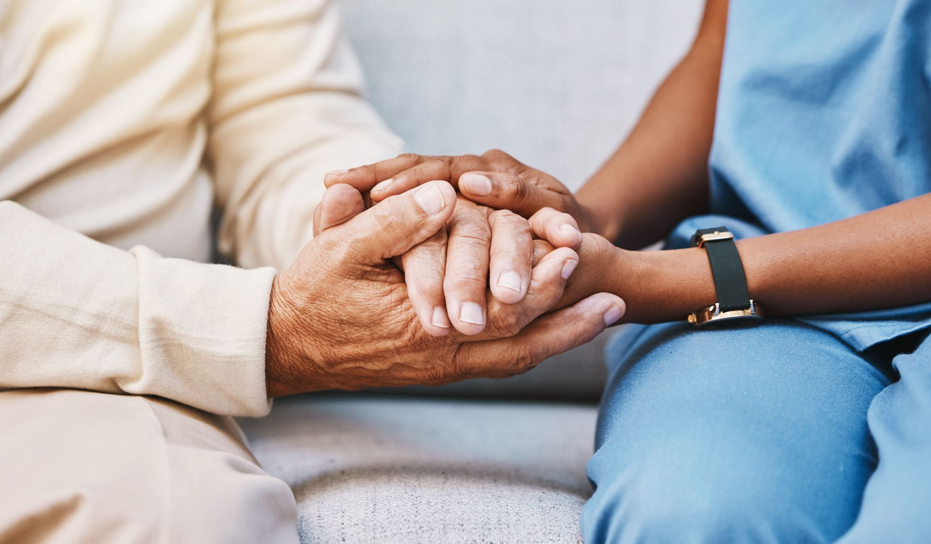 Two people holding hands on a couch, one in a cream sweater and one in blue scrubs with a watch.
