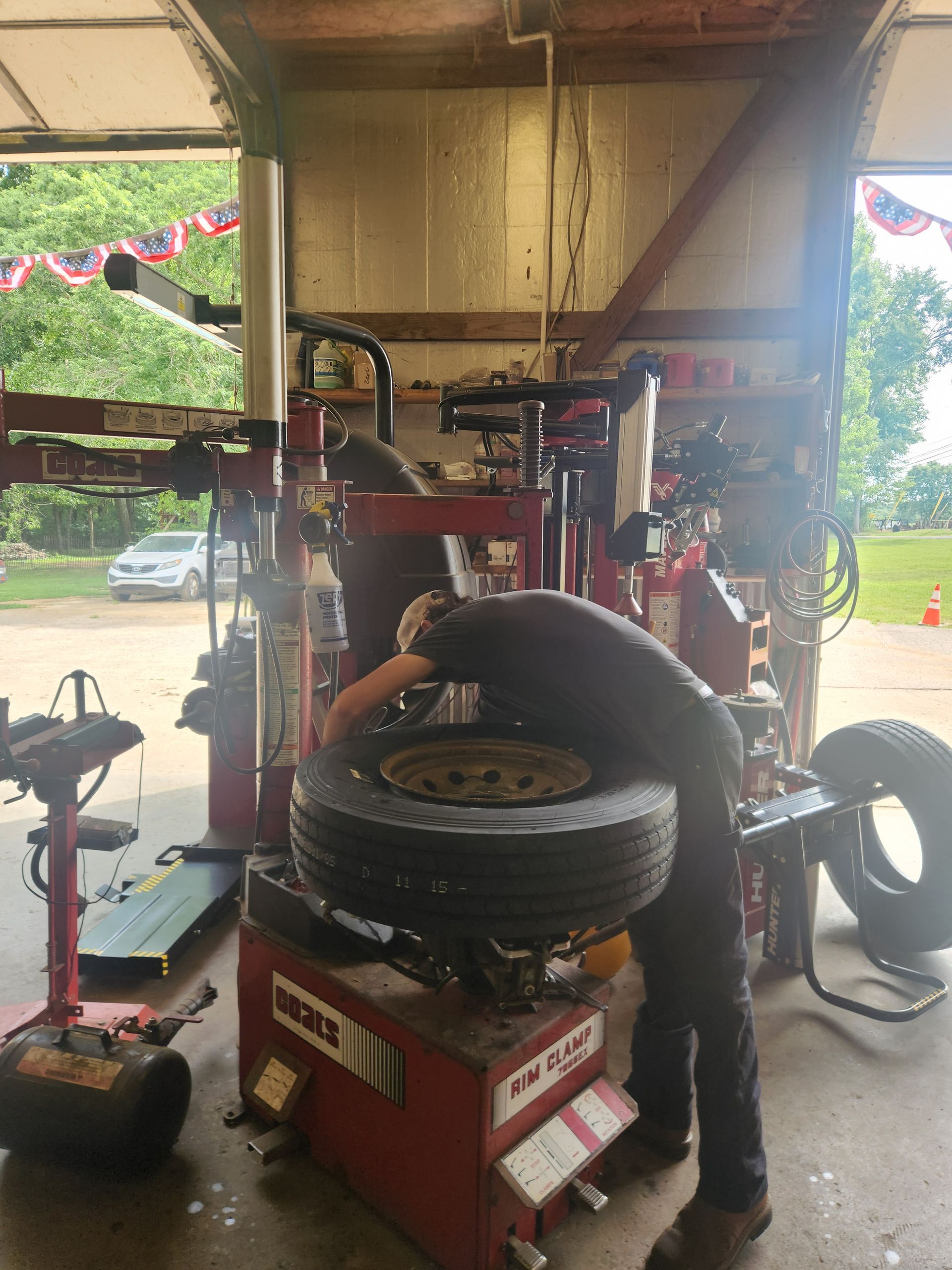 Mechanic bending over a tire-changing machine in a garage, with stacked tires and tools nearby