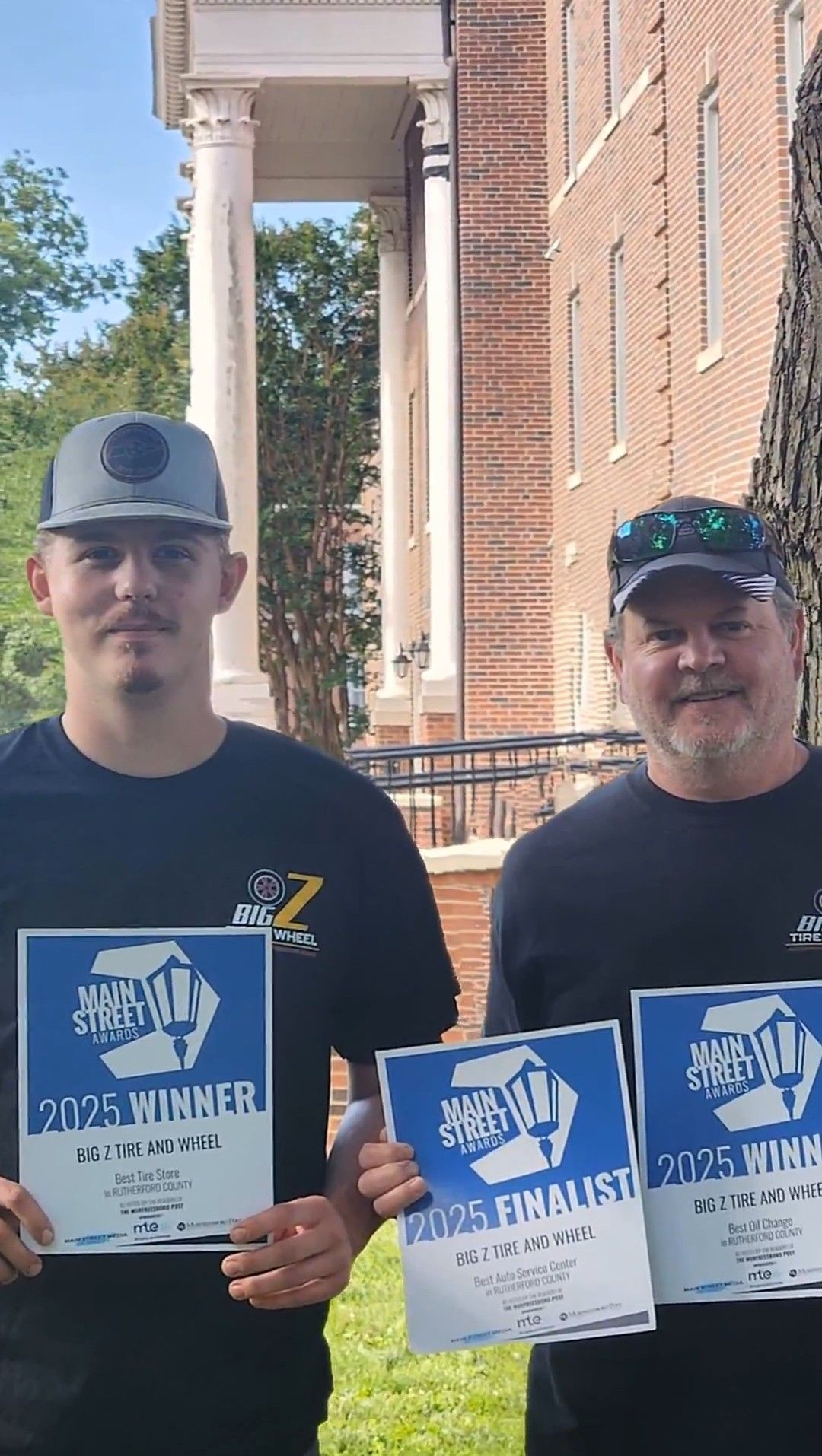 Two men holding blue certificates and posing outside a brick building with white columns.