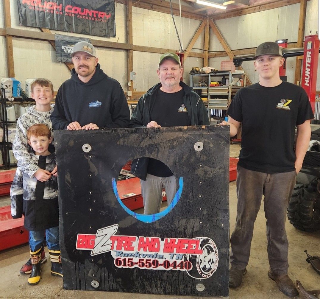 Four people holding a black fire pit sign in a workshop, with tools and tires in the background.