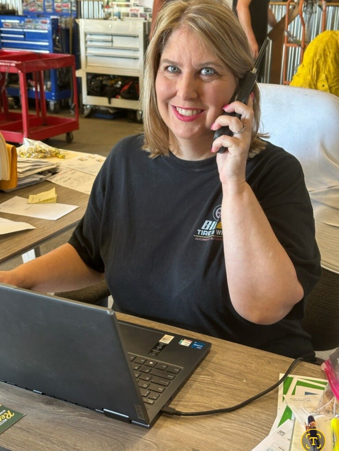 Woman smiling on the phone at a desk with a laptop in a busy workspace
