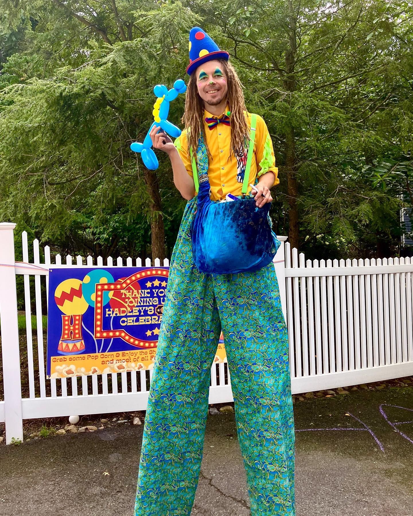 A man in a clown costume is standing on stilts holding balloons.