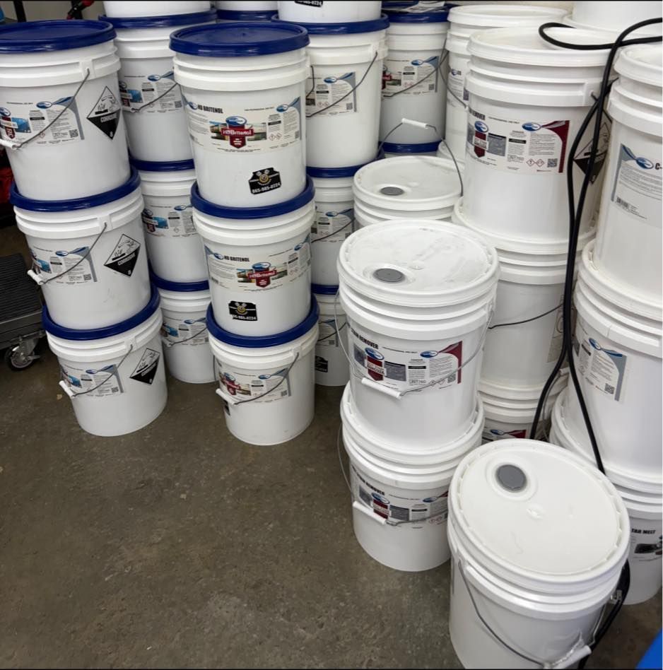 White buckets stacked, some with blue lids, containing a chemical substance. Hazardous material label visible.
