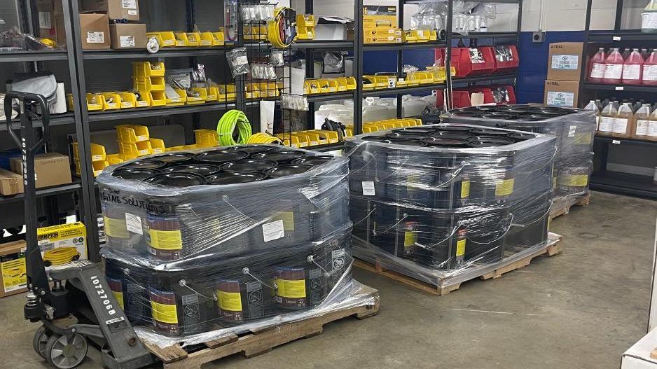 Pallets of black containers wrapped in plastic, on a warehouse floor. A forklift is to the left.