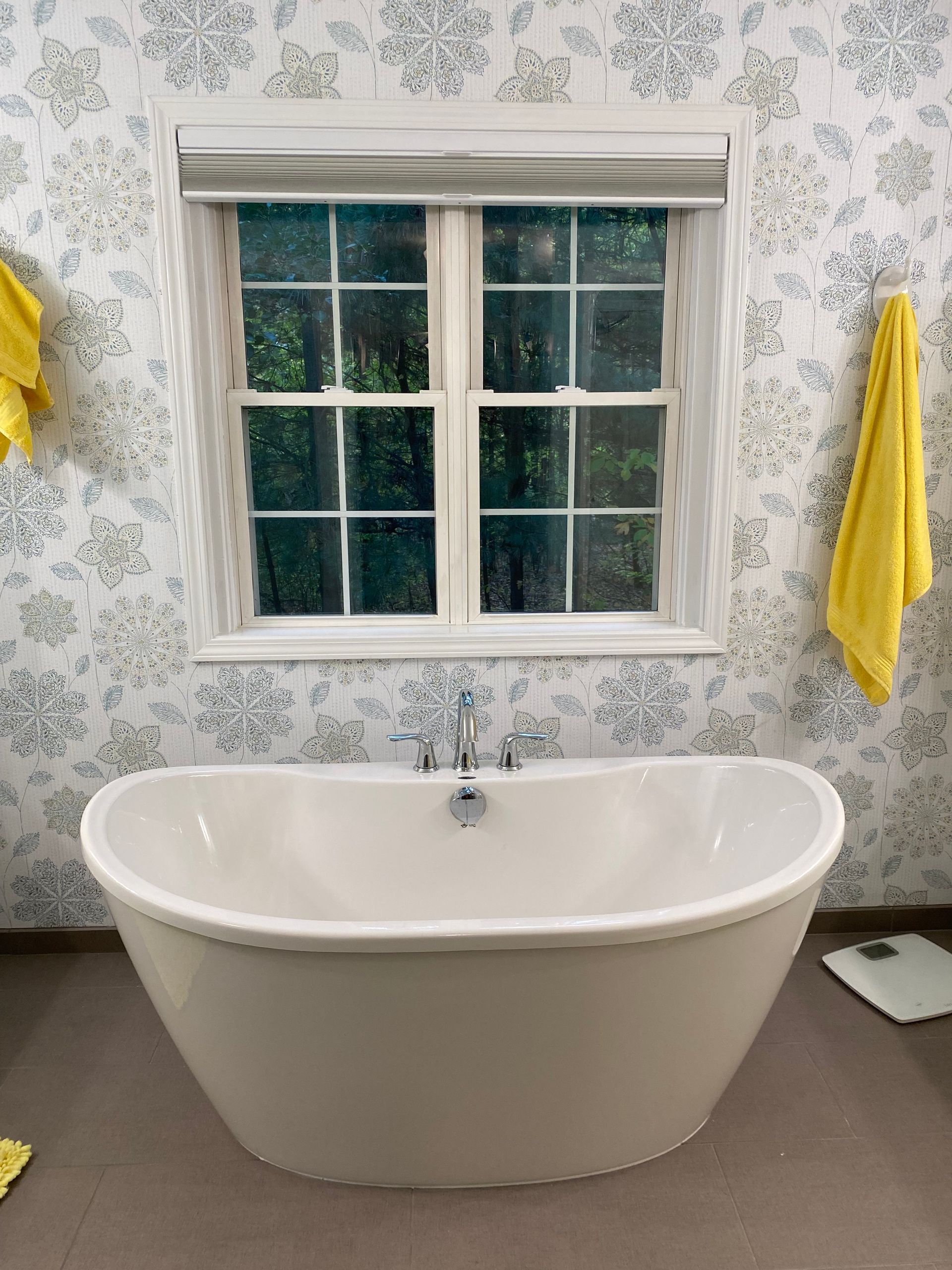 Freestanding bathtub in bathroom with floral wallpaper, window, and yellow towels.
