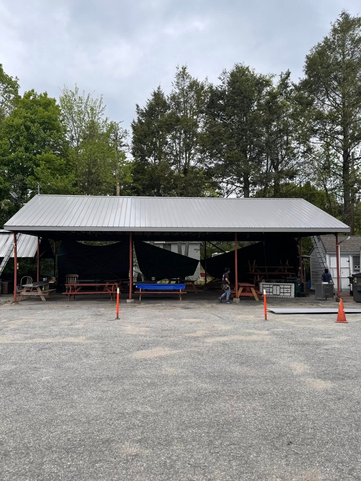 A covered outdoor eating area with picnic tables, a metal roof, and a gravel ground. The sky is overcast.