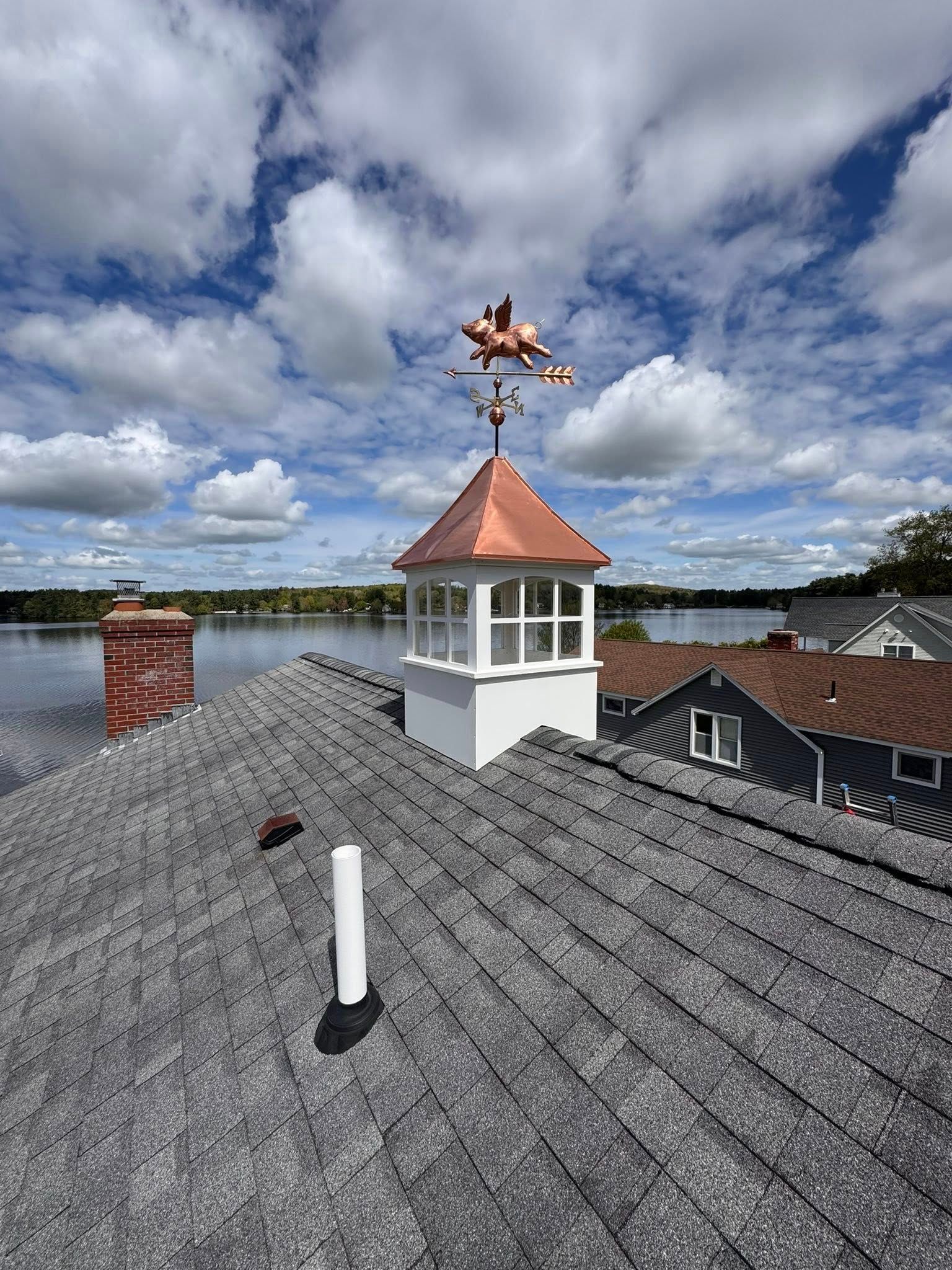 View of a rooftop with a white cupola topped with a copper roof and weather vane depicting a biplane; lake and sky in the background.