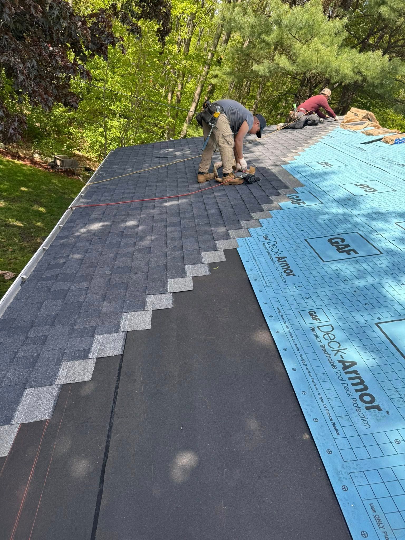 Roofers installing asphalt shingles on a house roof. One is kneeling, using a tool, while another works in the background.