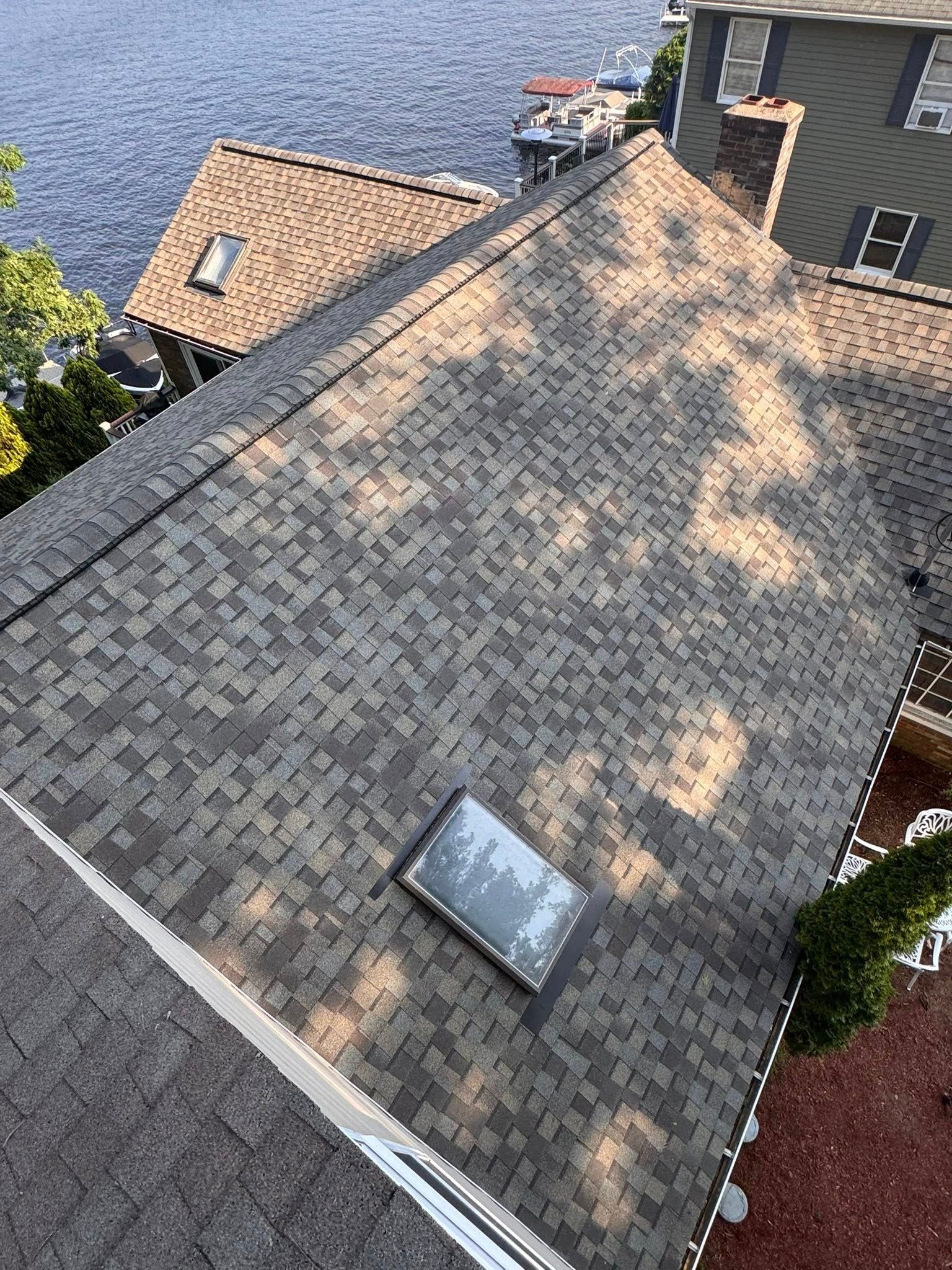 A shingled roof with a skylight and surrounding trees near a body of water. Overhead view, sunny day.