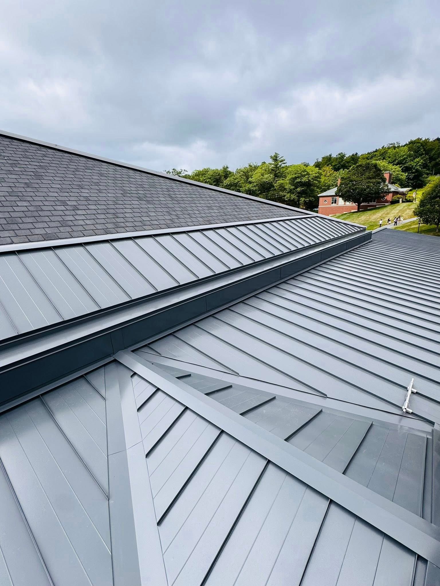 A gray metal roof of a building against a cloudy sky, with trees in the background.