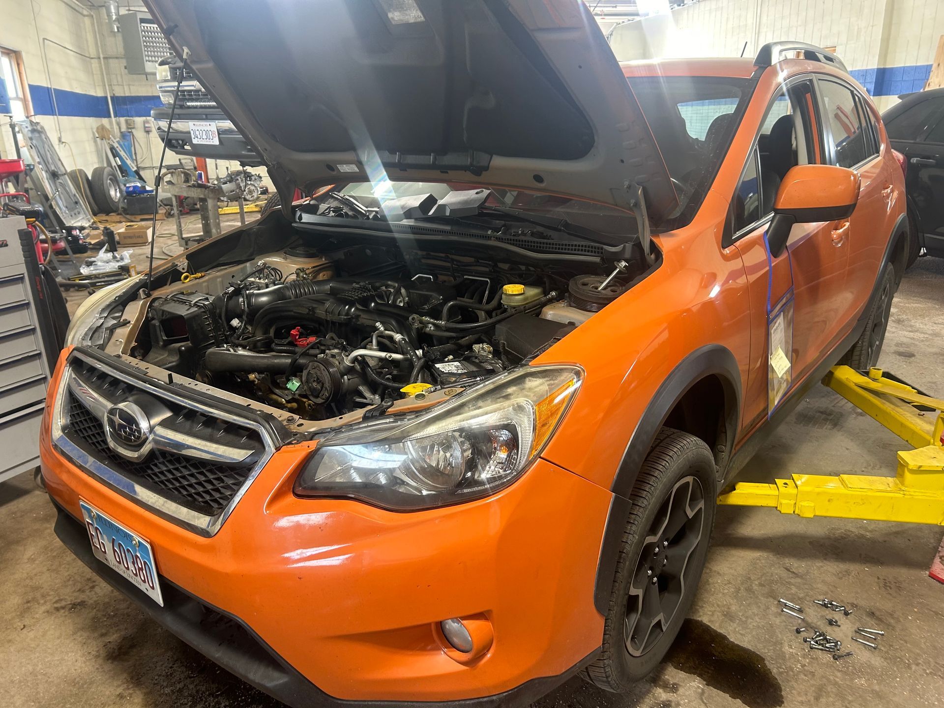 An orange subaru with the hood up is on a lift in a garage.