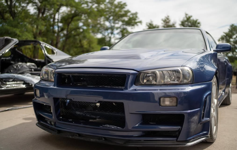 Blue Nissan Skyline R34 sports car with a modified front bumper, parked outside.