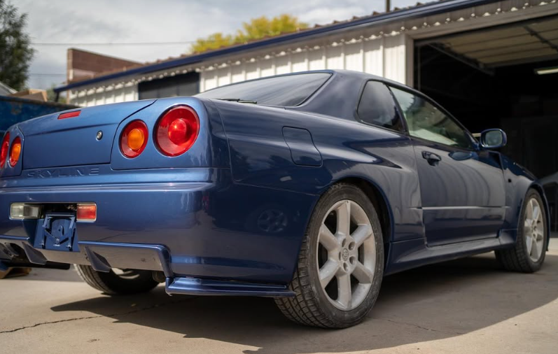 Blue Nissan Skyline R34 coupe parked outside a garage, rear view.