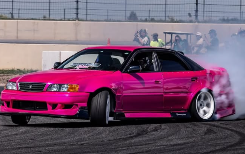 A hot pink drift car smoking its tires around a track. Spectators watch in the background.