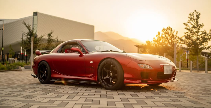 Red Mazda RX-7 sports car parked on a brick-paved area with mountains in the background, bathed in golden sunset light.