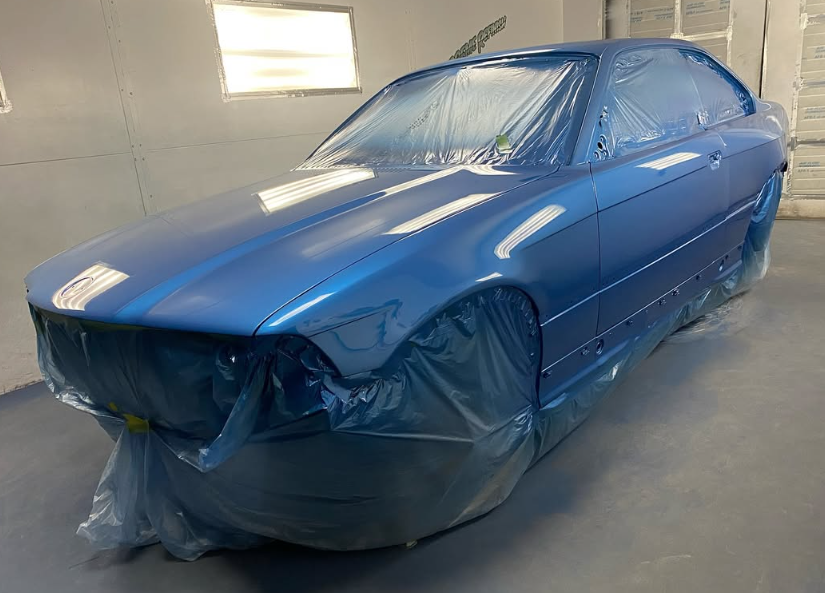 Blue car in a paint booth, covered in plastic sheeting.