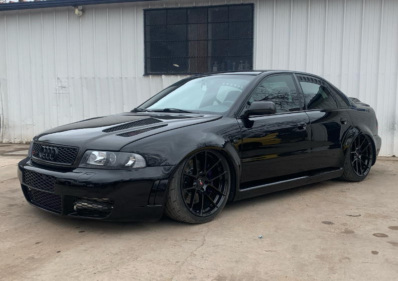 Black Audi sedan, lowered, with black wheels and a custom front grille, parked in front of a white building.