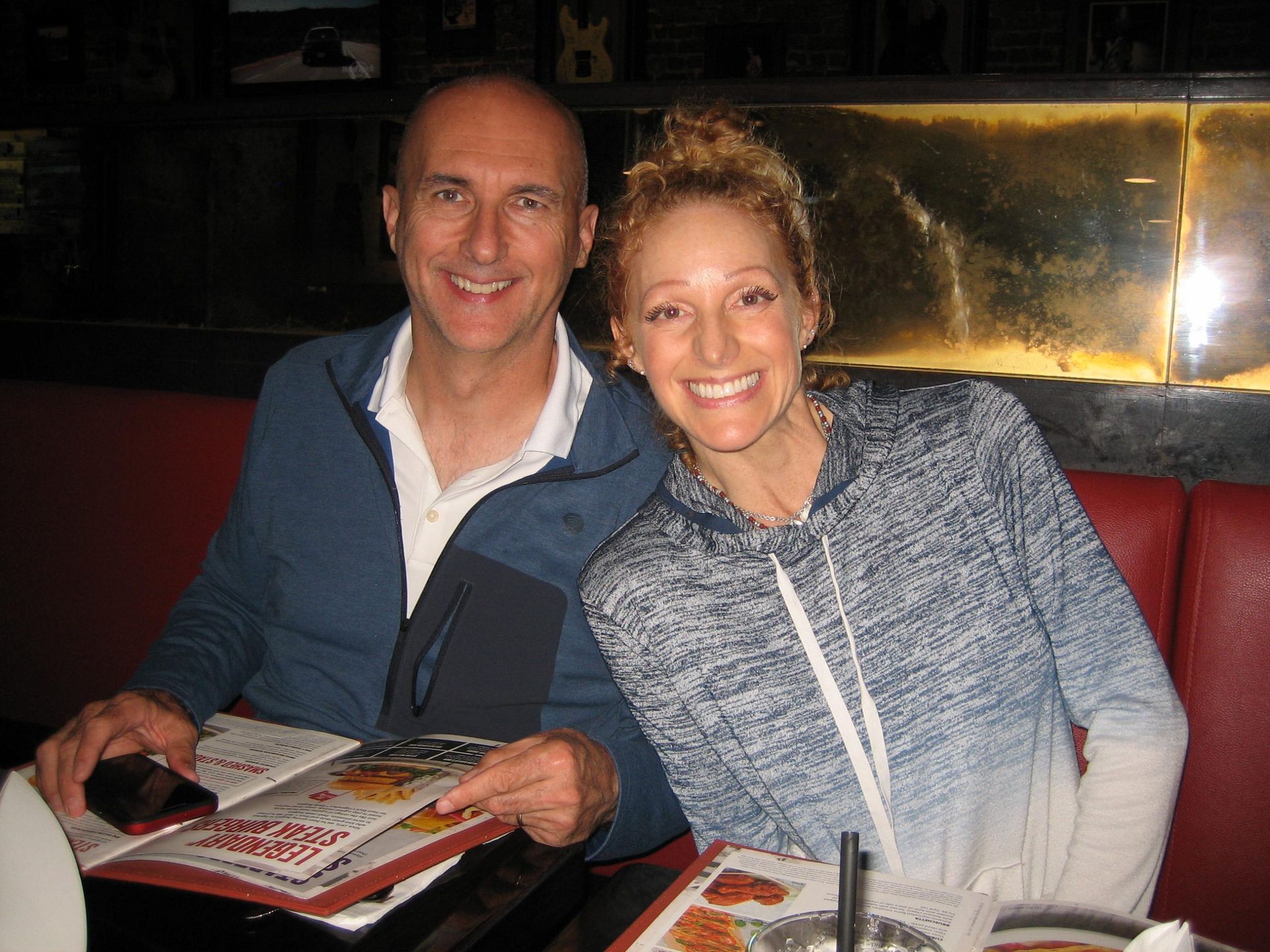 Two people smiling at a restaurant table, seated on a red booth with menus and drinks in front of them.