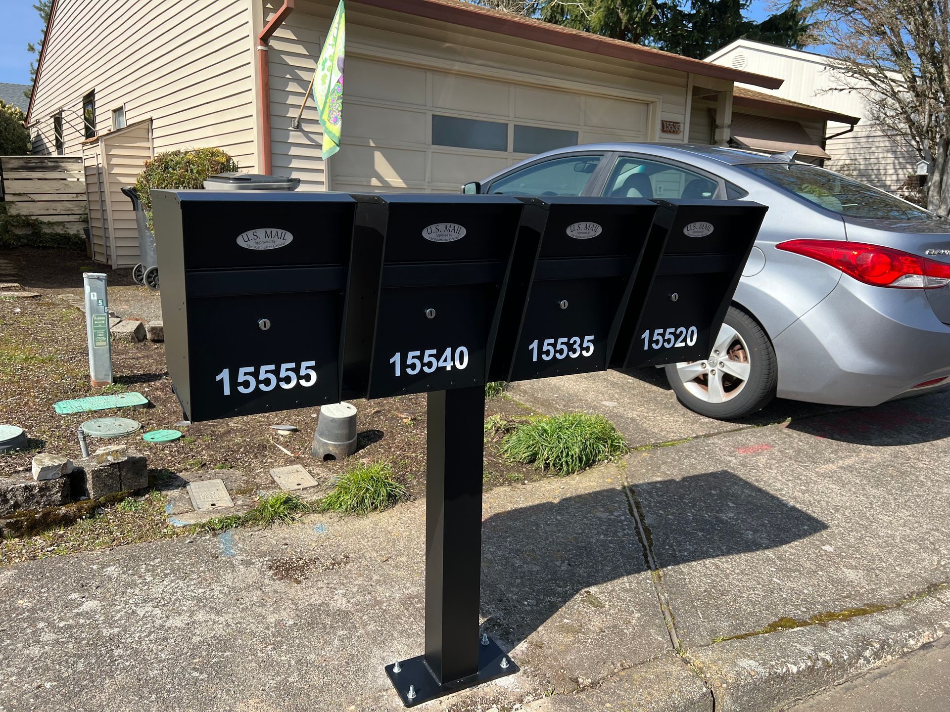 Four black mailboxes on a post in front of a house, with a silver car parked beside them.