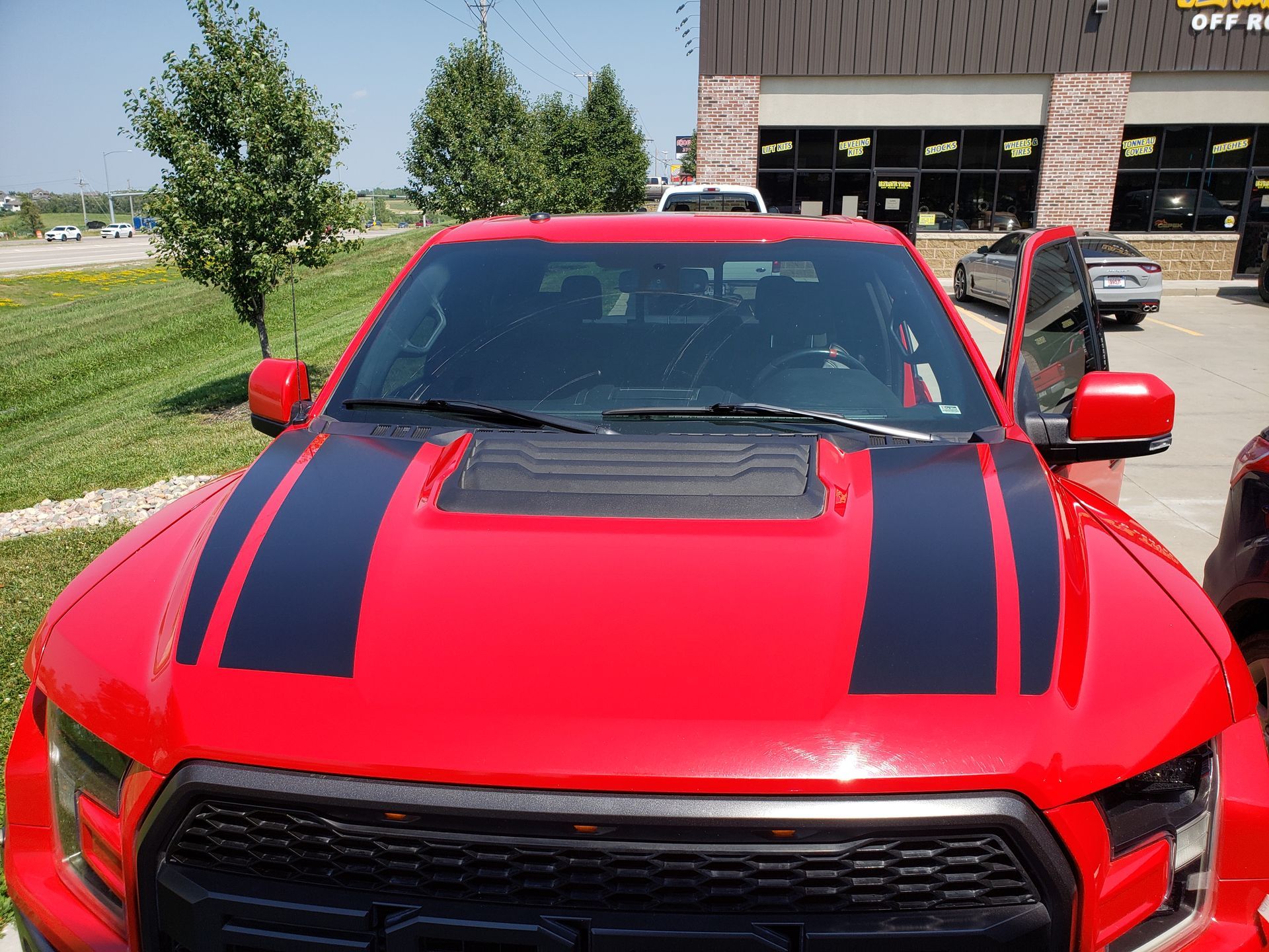 Red Ford Raptor truck with black stripes on hood, parked outside a building.