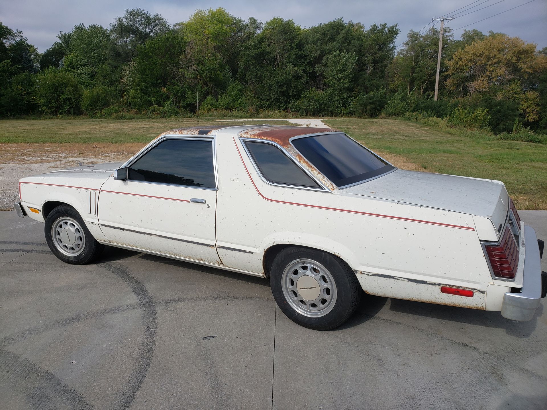 White 1980s Ford Fairmont Futura coupe with rusty roof, parked on concrete. Trees in background.