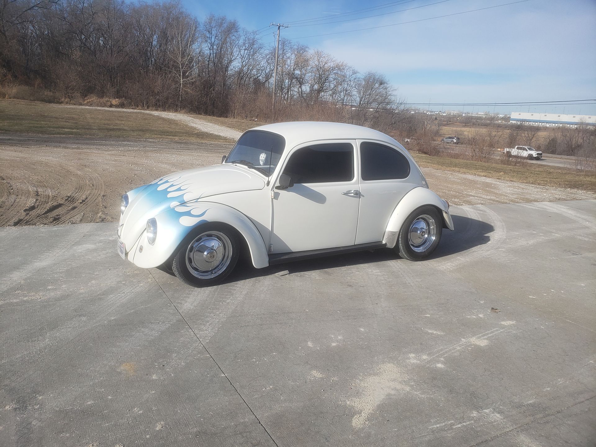 White Volkswagen Beetle with blue flame decals parked on a concrete surface, with a natural background.