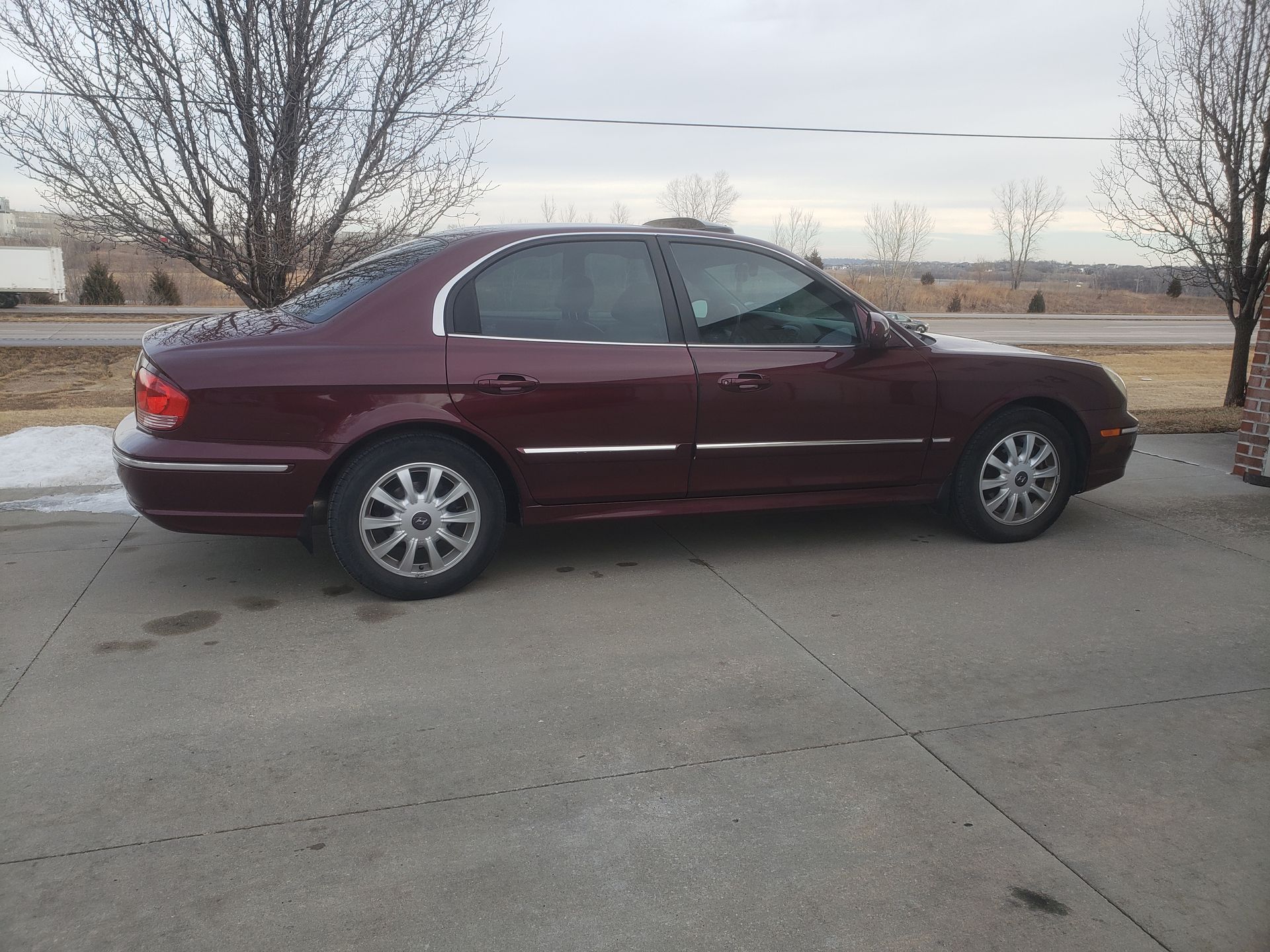Maroon sedan parked on concrete, near trees and a building in winter.