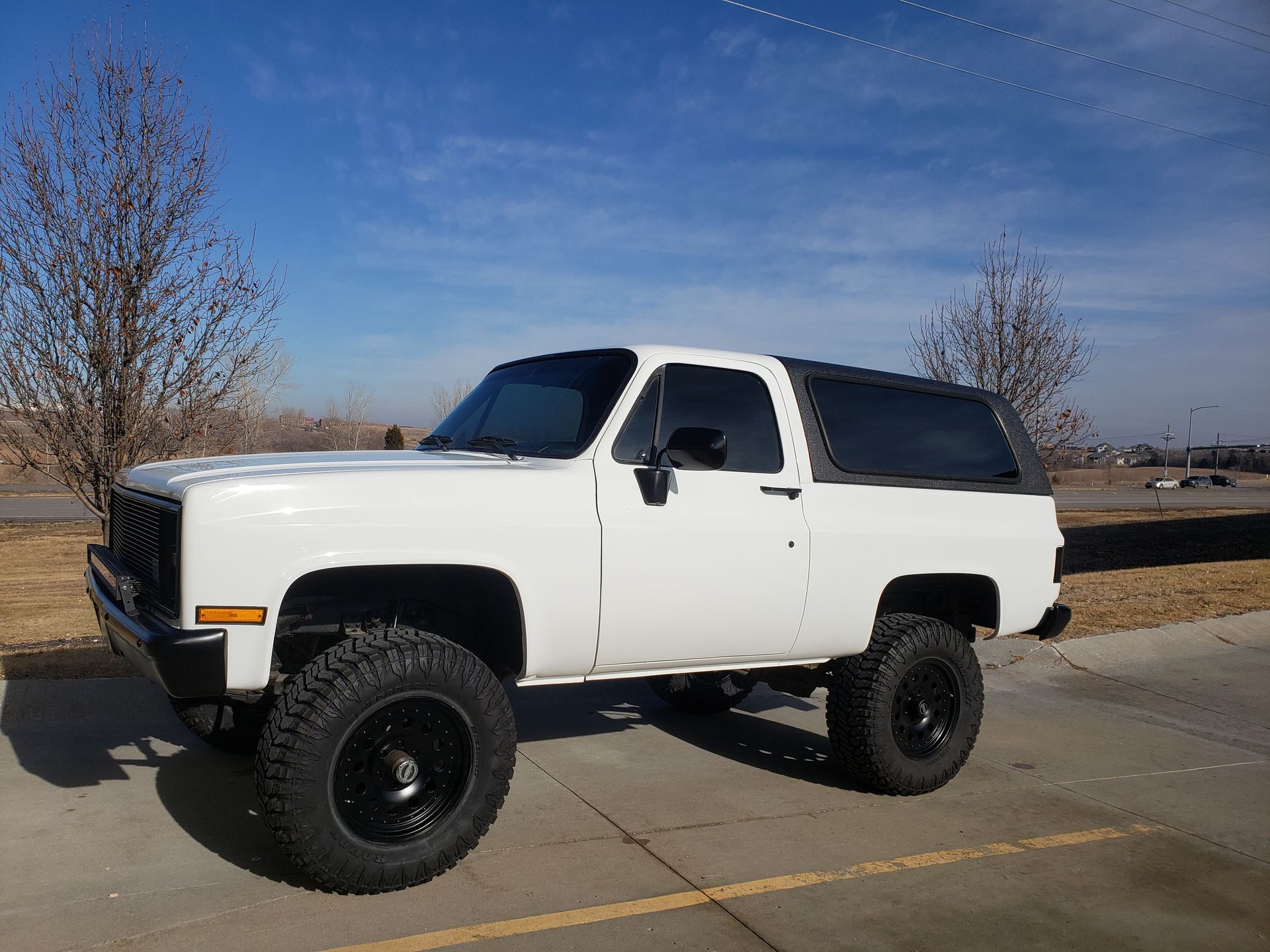 White, lifted SUV with black wheels and a black top parked on pavement under a blue sky.