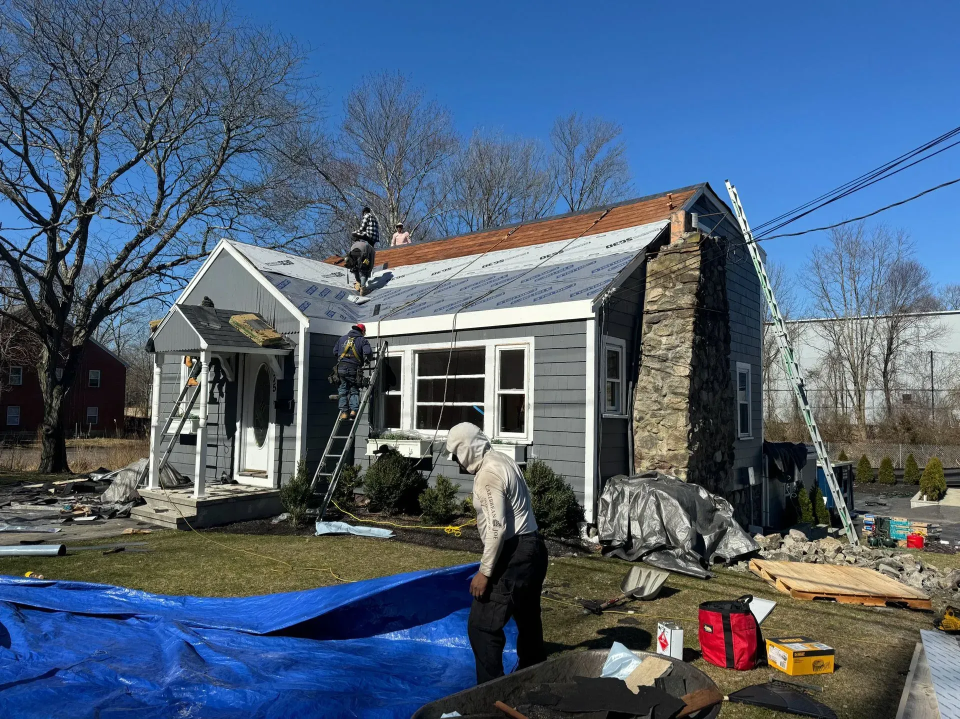 Roofers working on a house with a gray exterior and a stone chimney on a sunny day.