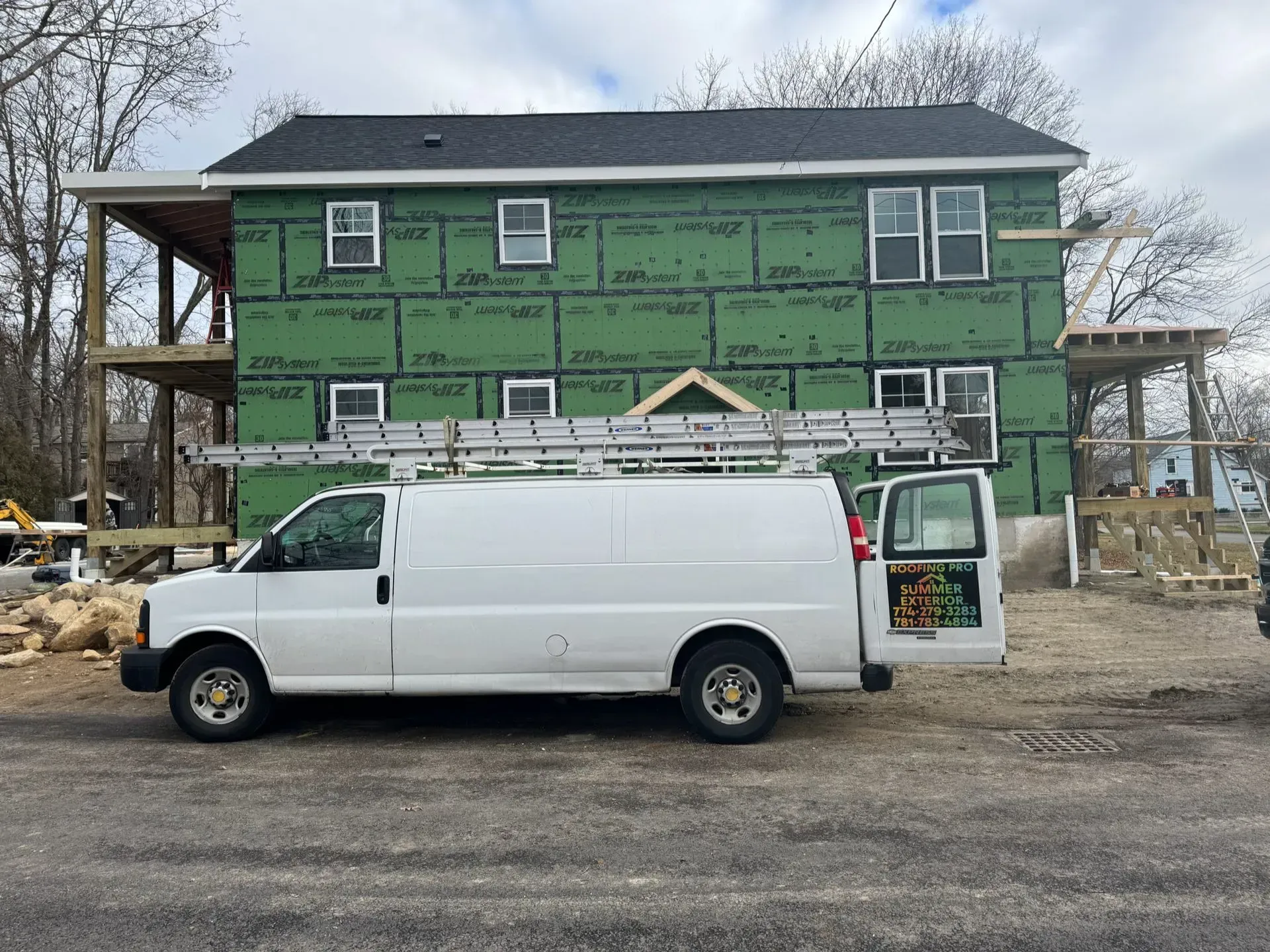 White work van with ladder parked in front of a house under construction. Exterior walls are green. Cloudy day.