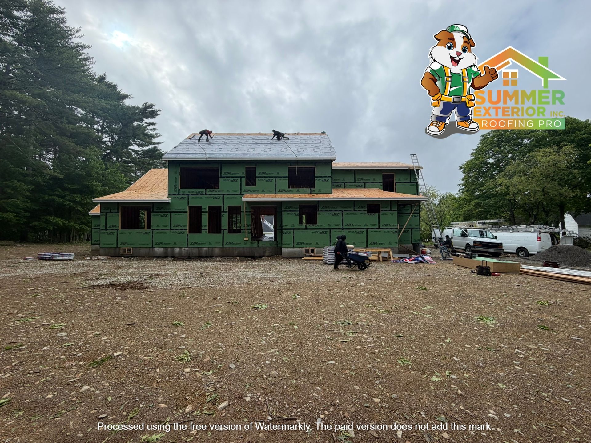 Green two-story building under construction in a dirt clearing, with trees and construction materials nearby.