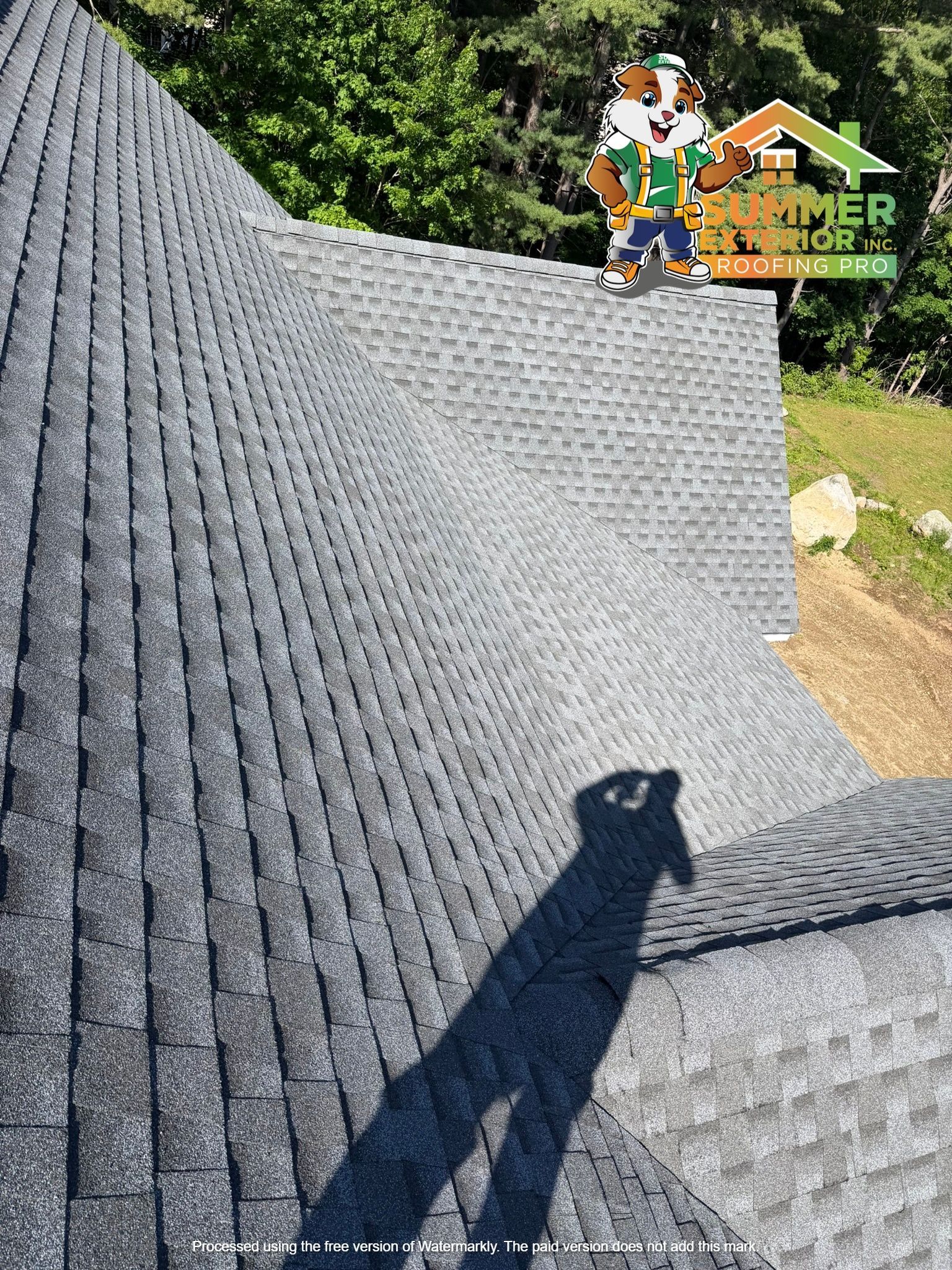 Shingled roof with a person’s shadow in the foreground and a worker standing on the ridge near trees and a sign.