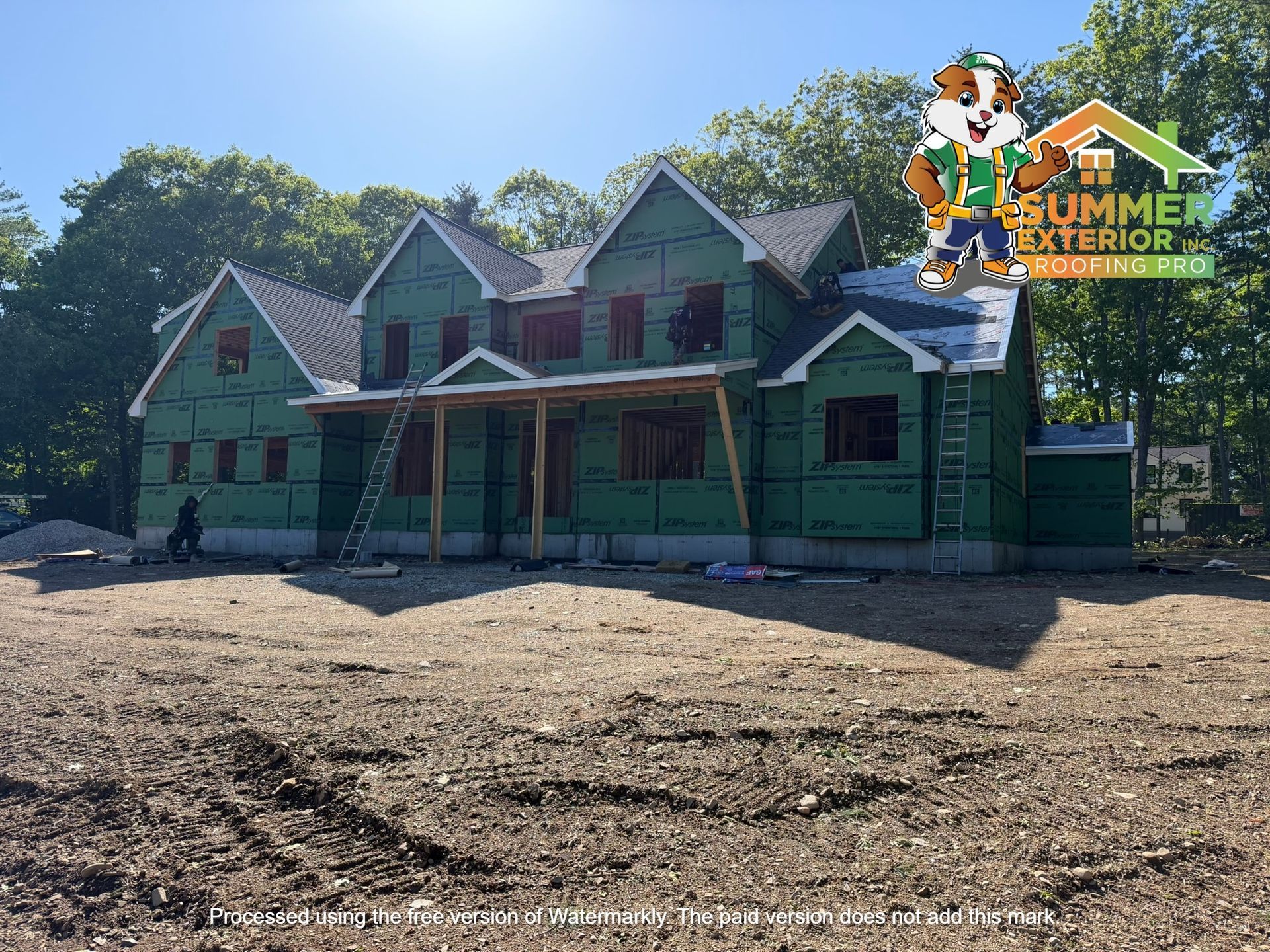 Green house under construction with teal siding and a front porch on a dirt lot, under a clear blue sky
