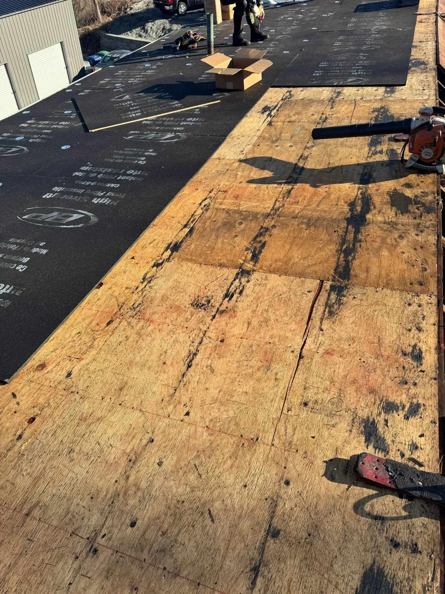 Roof partially stripped of shingles, showing exposed wood, tools, and a person in the background.