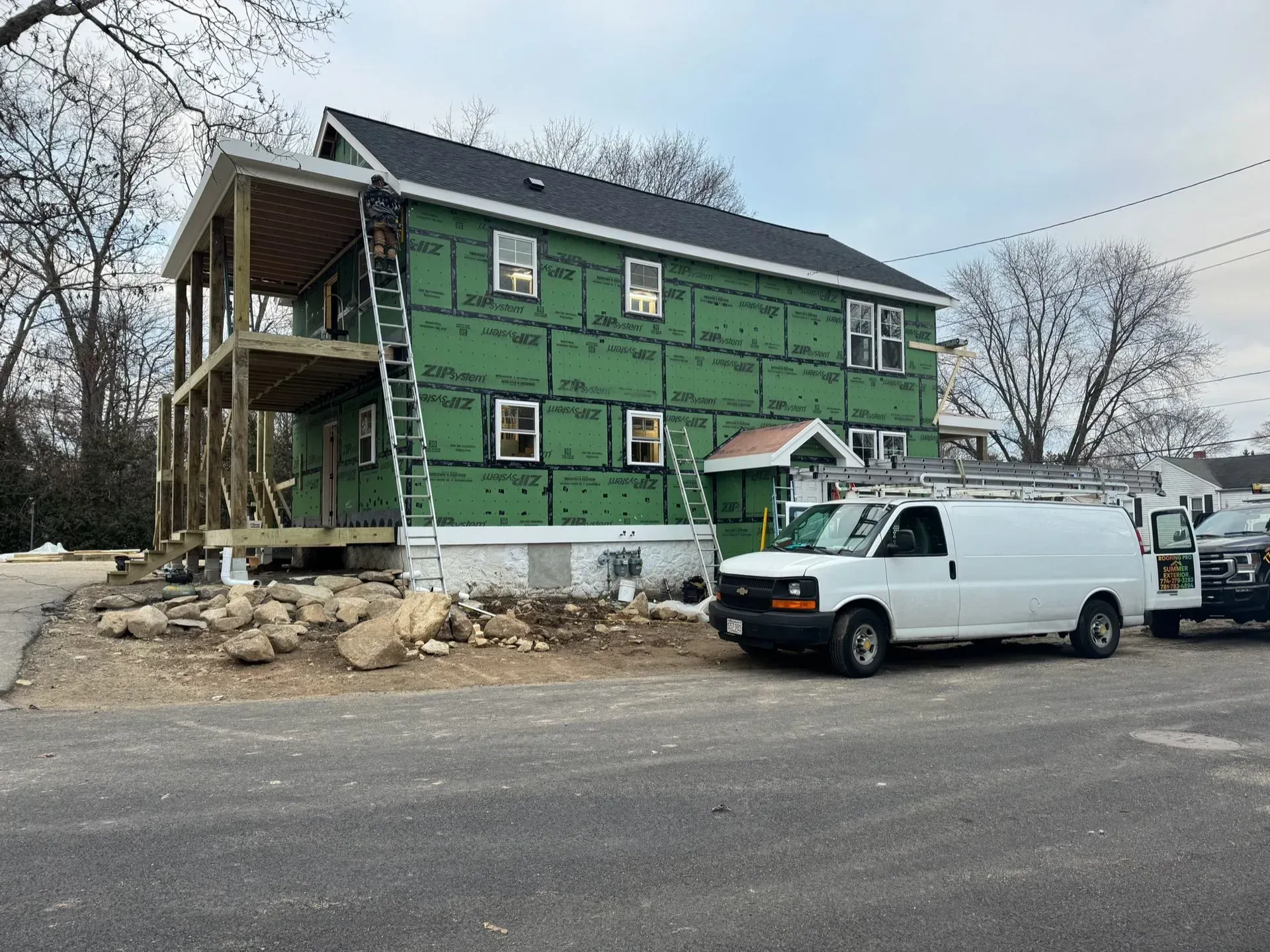 Two-story house under construction, green sheathing, white vans parked nearby, ladder leaning against the house.
