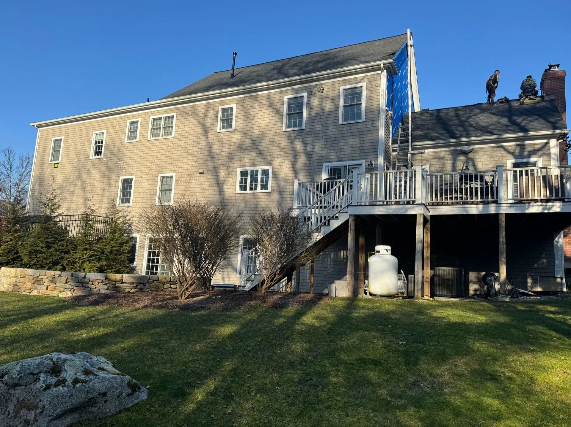 Two-story house with workers on the roof. Deck with a propane tank, and a grassy yard in the foreground.