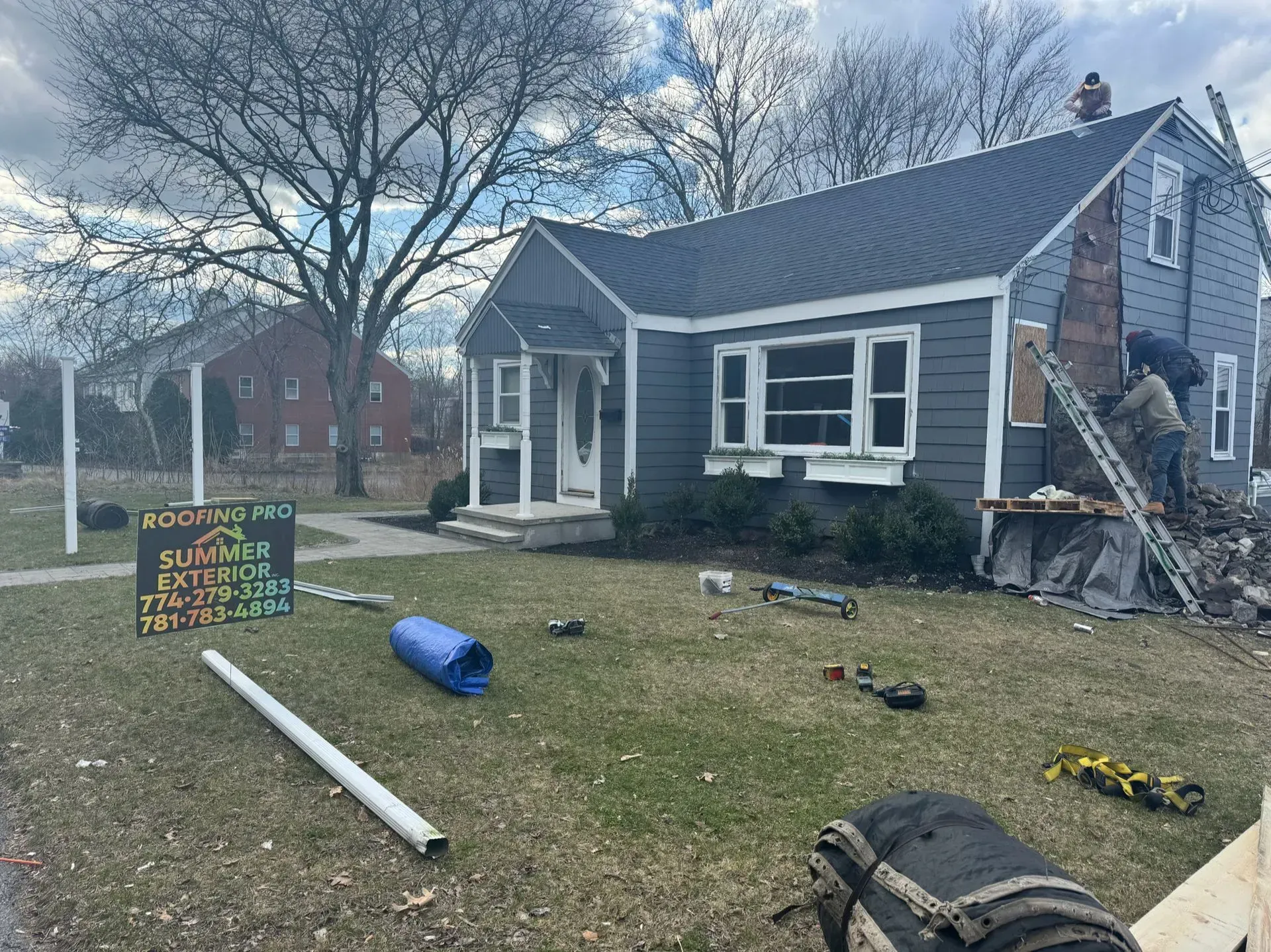 House with dark gray siding, roof repair in progress. Workers on ladder near a brick chimney.