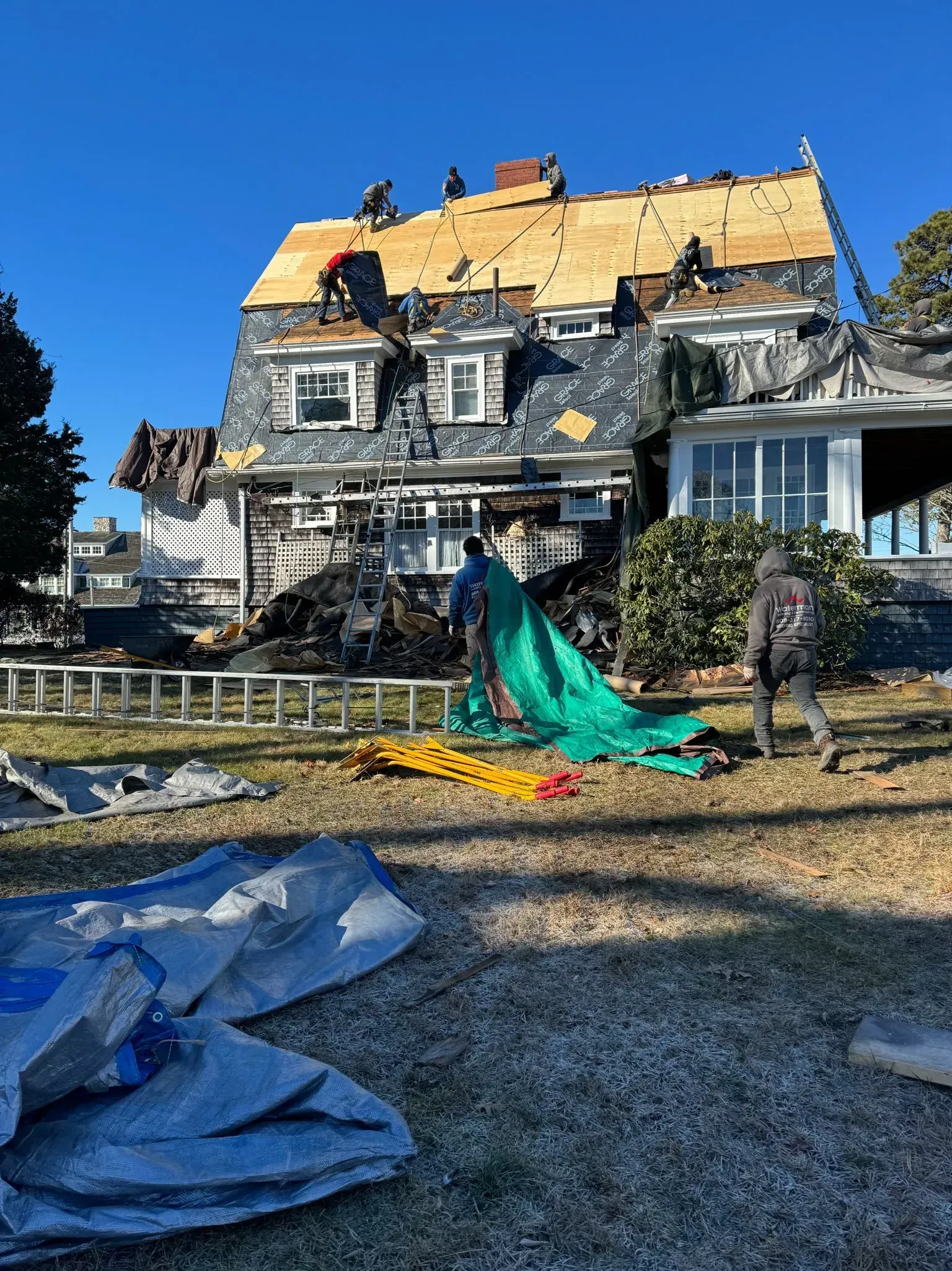 Roof being replaced on a two-story house; workers on roof, green tarp on ground, clear sky.