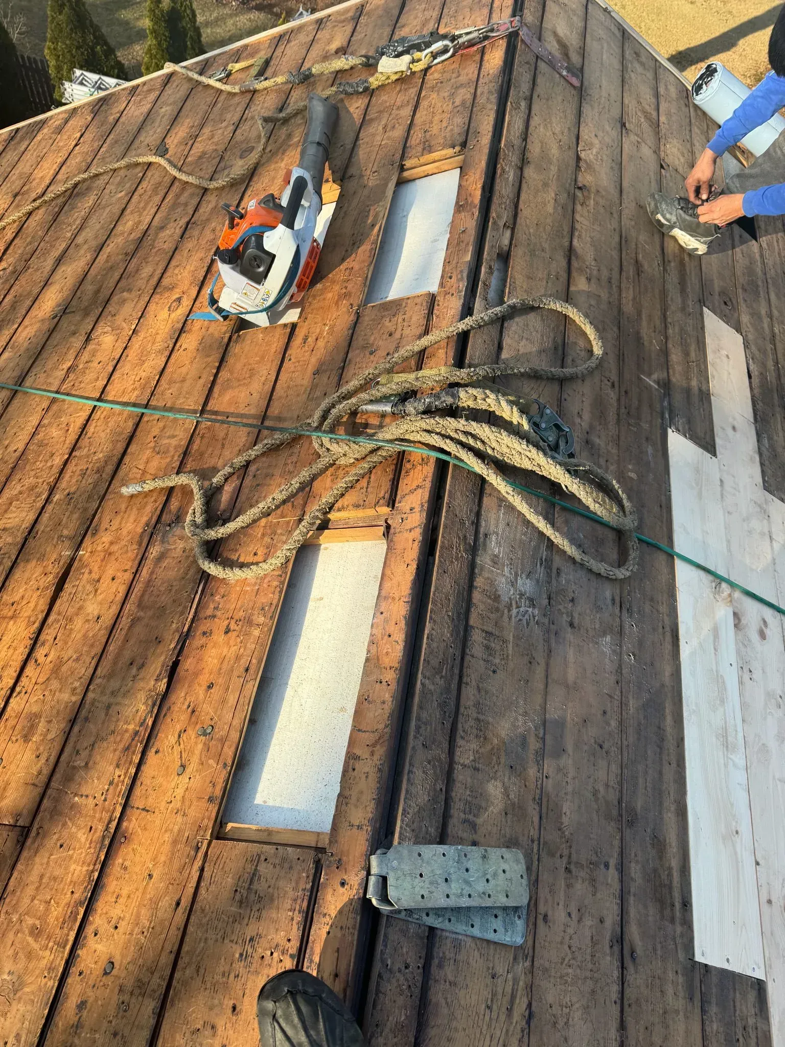 Roof with wooden planks, holes cut for skylights. Tools and rope present. Person working.