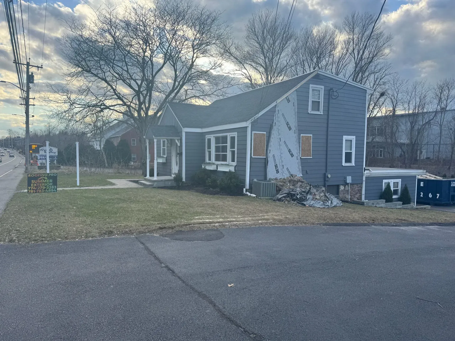 A blue house with missing siding is under construction on a grassy lot, viewed from a street.