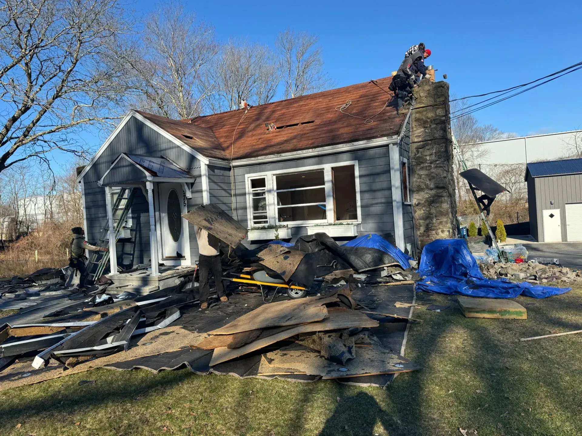 House with damaged roof and debris; two workers on roof, blue tarp in yard.