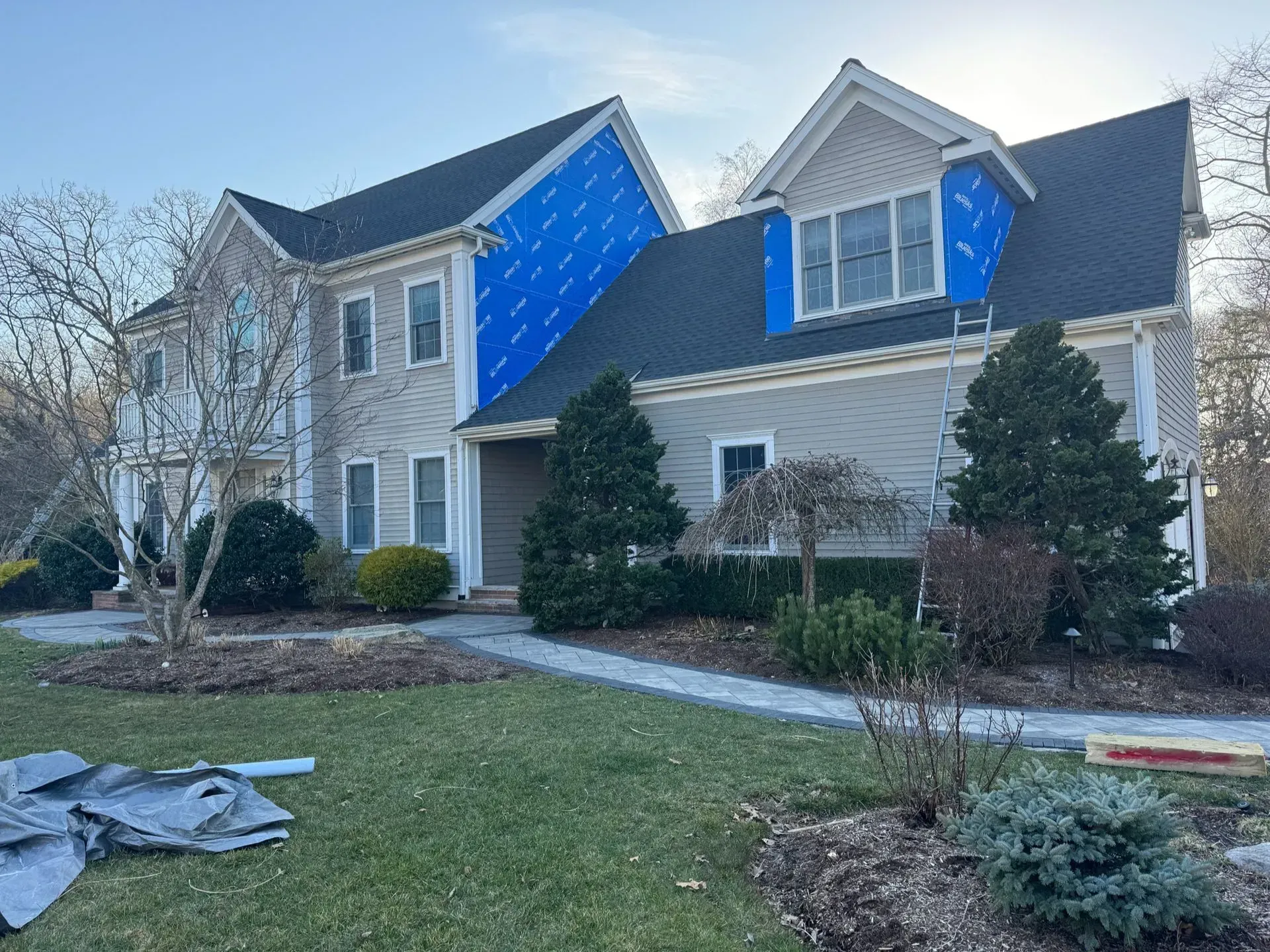 Two-story house undergoing exterior renovation; blue protective covering on the roof, gray siding, and green lawn.